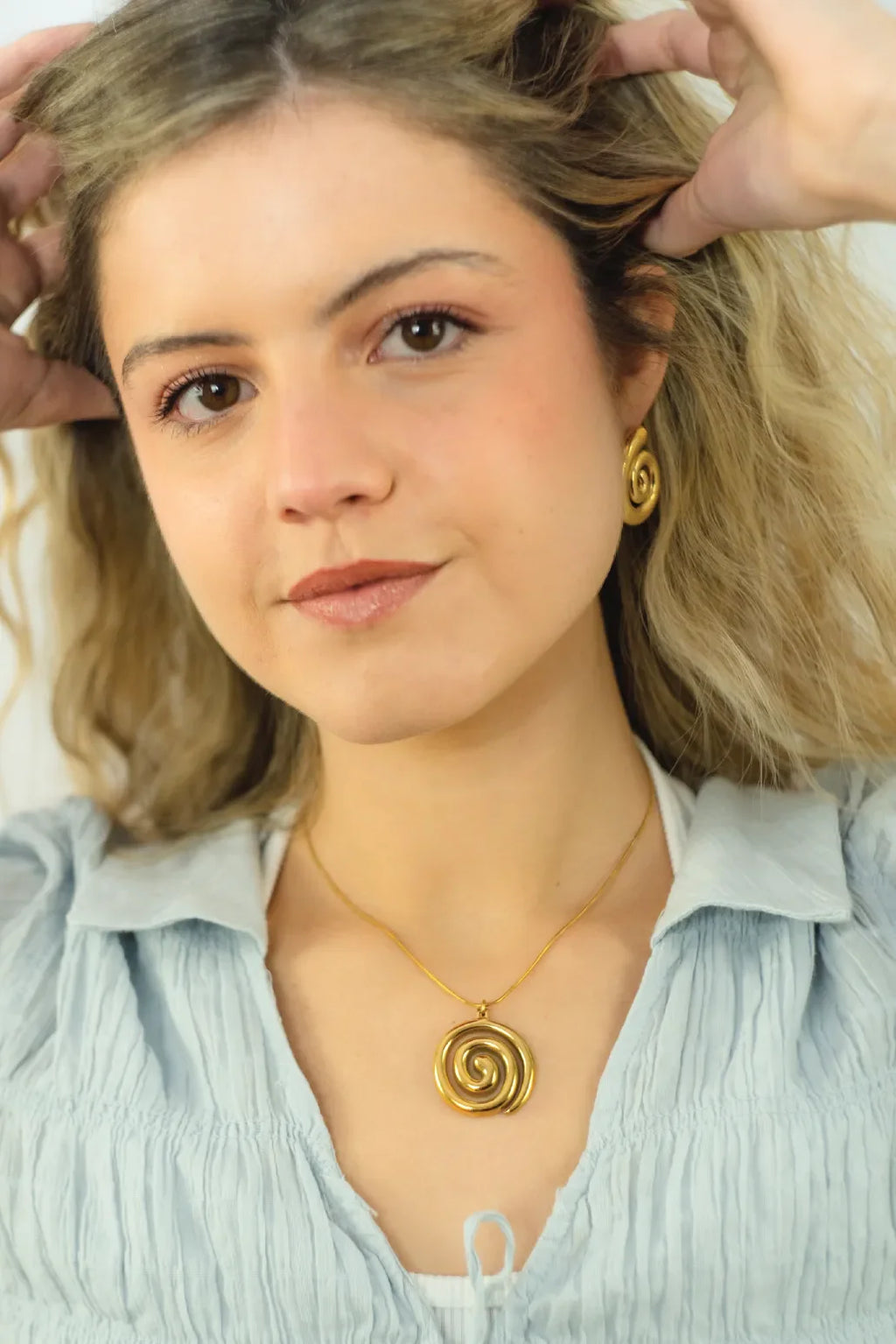 Woman wearing a gold spiral necklace and earrings, with hands adjusting her hair.